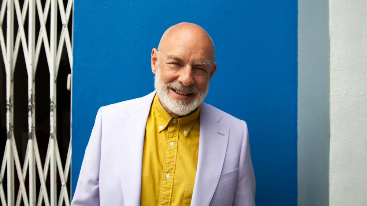 Brian Eno in a pale jacket and yellow shirt against a blue wall.