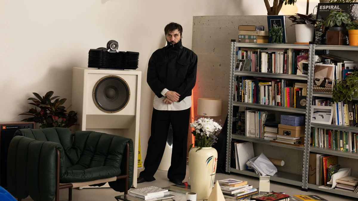 John Talabot standing beside a speaker and shelves of records and books.
