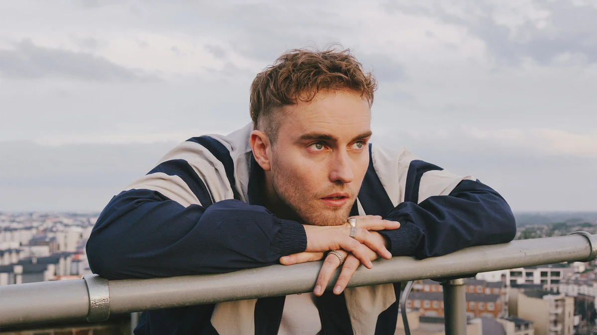 Sam Fender leaning on a rooftop railing with a city skyline behind him.